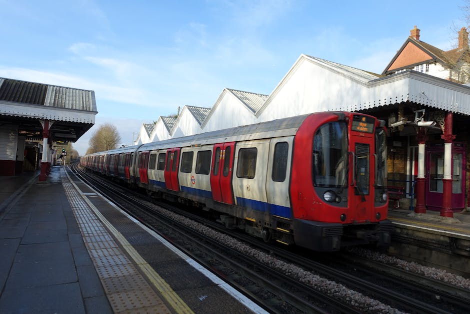 A busy street scene outside St Johns Wood Underground Station in London, with pedestrians walking on the pavement and some carrying luggage or bags. In the foreground, a row of metal bollards lines th